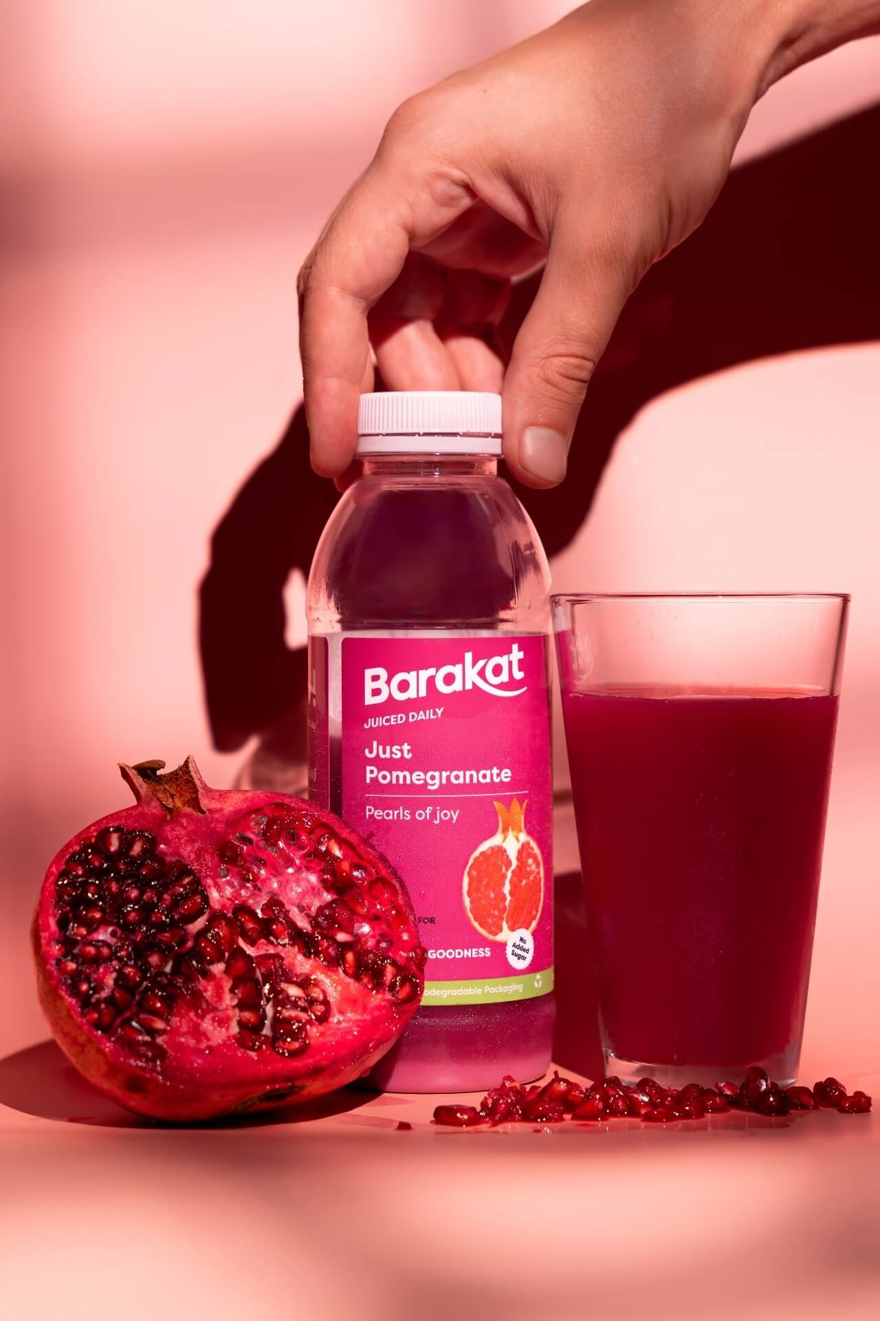 Hand of a man holding a Barakat Pomegranate bottle next to a glass with juice and a pomegranate